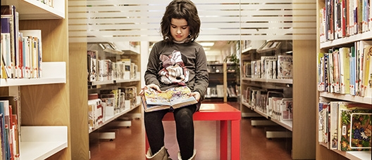 Niña sentada sobre una mesa leyendo un libro en un pasillo de la biblioteca