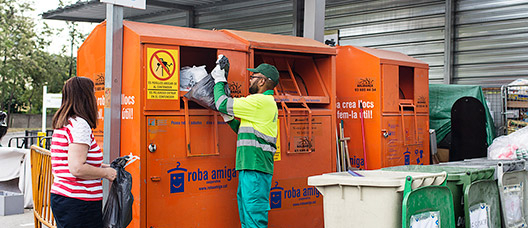 Un treballador de la deixalleria atén una usuària que porta roba a un contenidor de reciclatge.