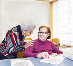 Dos usuarias de un centro de día hablando mientras una realiza una actividad de escritura