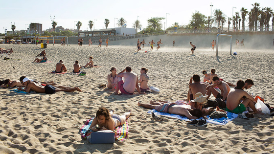 Vista de la arena de una playa con grupos de personas tumbadas y grupos de personas que practican deportes