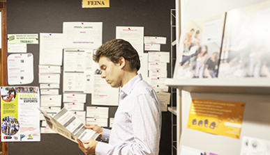 Home jove al costat d'un tauler d'anuncis de feina llegint una oferta de treball